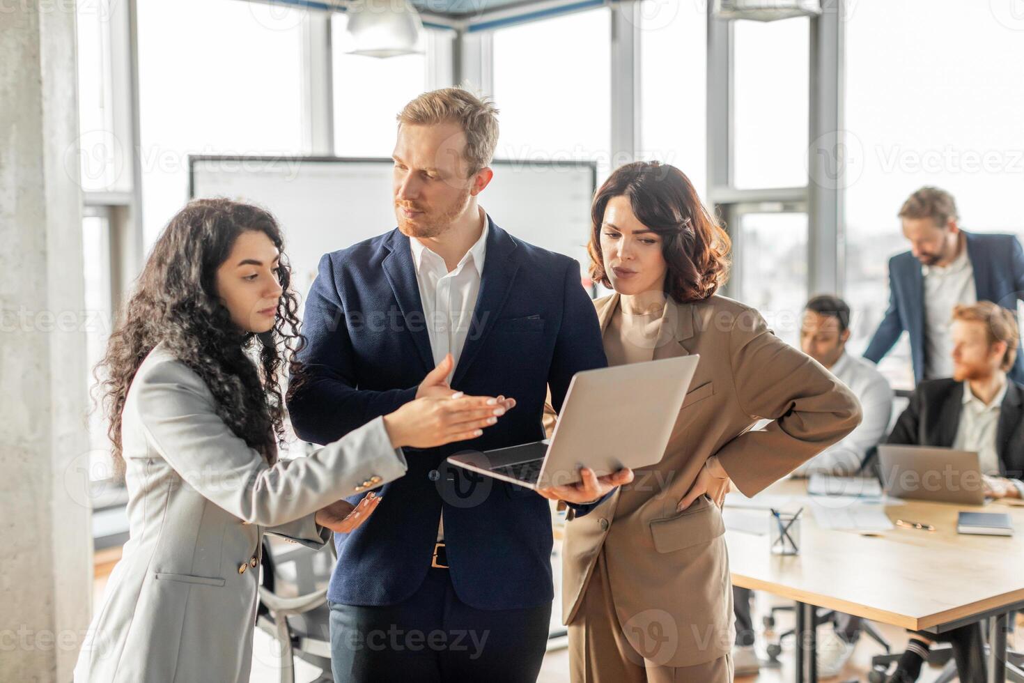 Three business professionals stand together in a modern office, engaged in a collaborative discussion while looking at a laptop. The man in the center appears to be leading the conversation photo