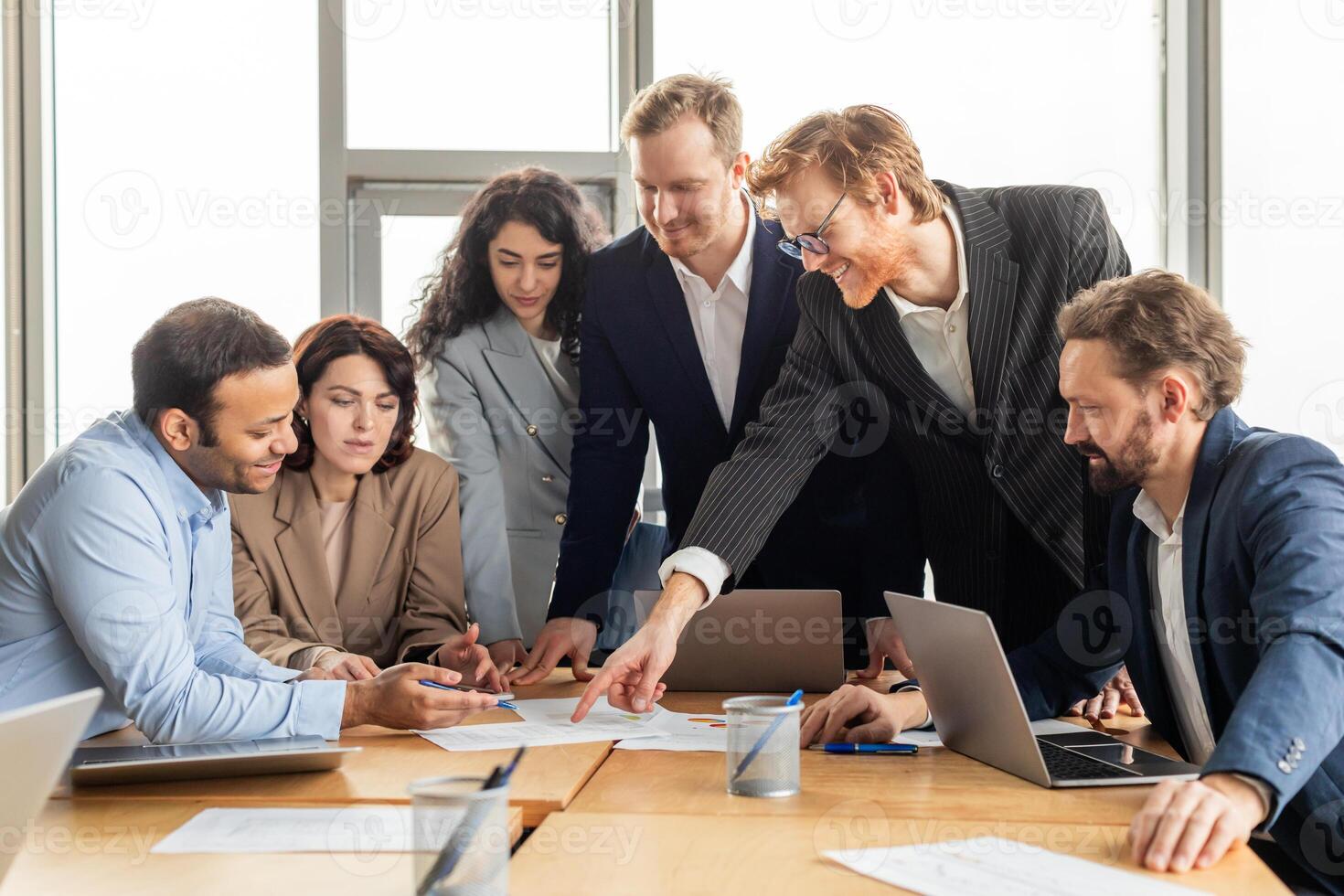 A group of business professionals are gathered around a table in a modern office, collaborating on a project. They are reviewing documents and using laptops. photo
