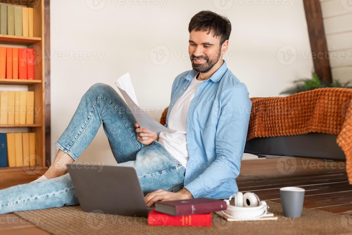 A cheerful man in casual attire sits on the floor with his laptop open and paperwork in hand, sitting area features a coffee cup, headphones, and books, suggesting a relaxing work-from-home setup photo