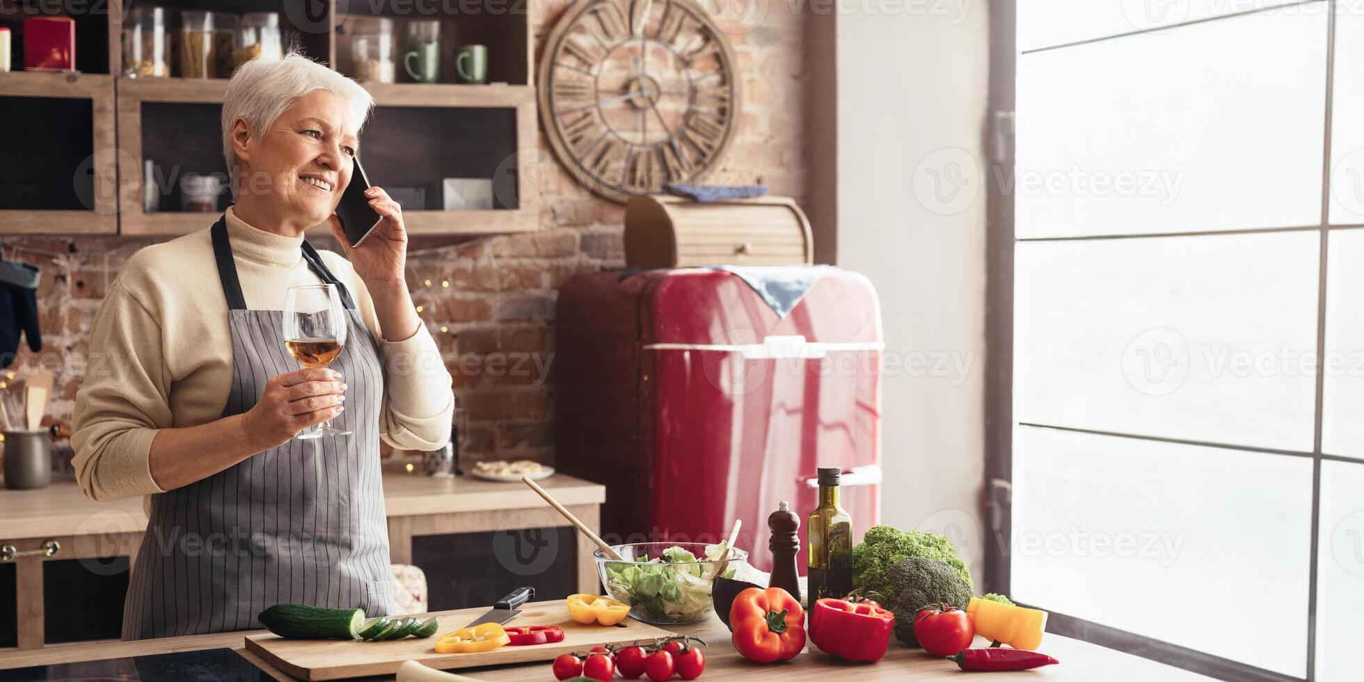 A senior woman stands in her modern kitchen, talking on her cell phone while holding a glass of wine. Fresh vegetables and a bowl of salad are on the counter, copy space photo