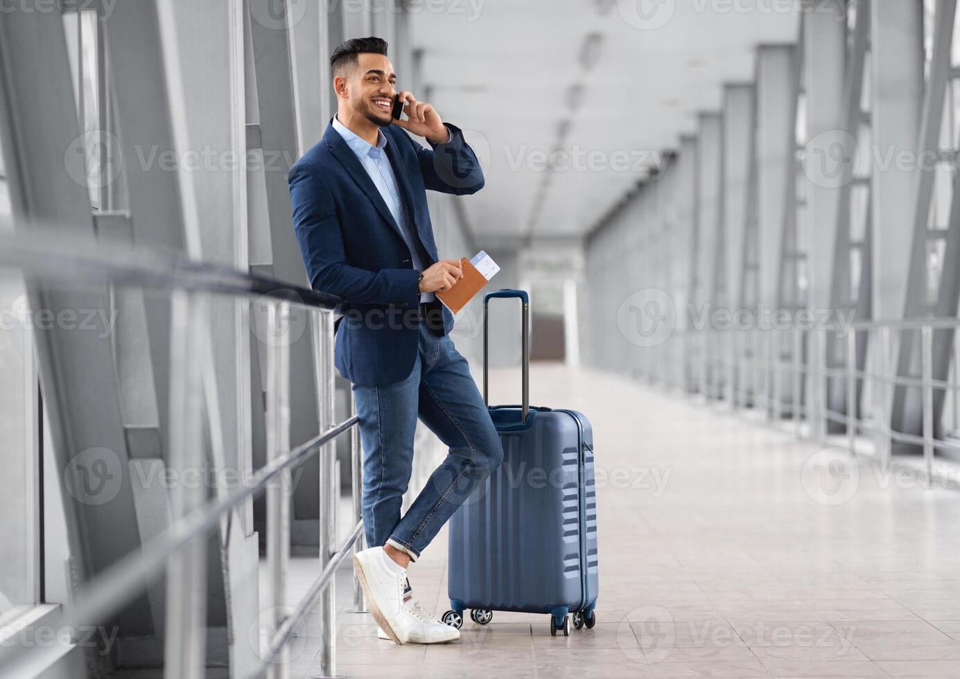 Mobile Call. Smiling Young Middle Eastern Businessman Talking On Cellphone In Airport While Waiting With Suitcase For Flight At Terminal, Happy Arab Man Having Pleasant Phone Conversation photo