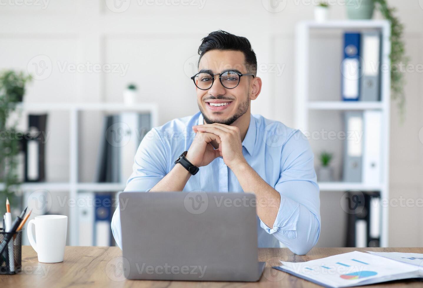 Portrait of professional young Arab guy sitting at his desk with laptop pc in modern office. Millennial businessman using computer for online job at workplace, having virtual meeting photo