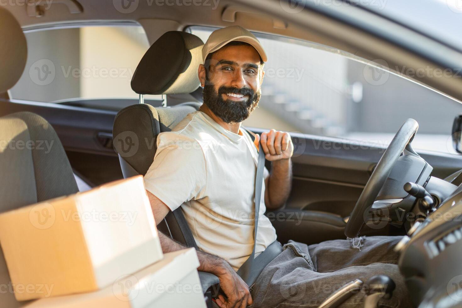 A bearded Indian man is sitting in the drivers seat of a car, smiling while fastening his seatbelt. Several packages are visible in the car. The background indicates a bright, sunny day. photo