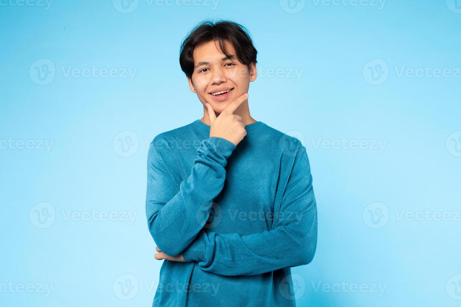 Asian young guy wearing a casual blue sweater places one hand on his chin, striking a thoughtful pose. He stands against a light blue background, creating a calming and serene atmosphere. photo