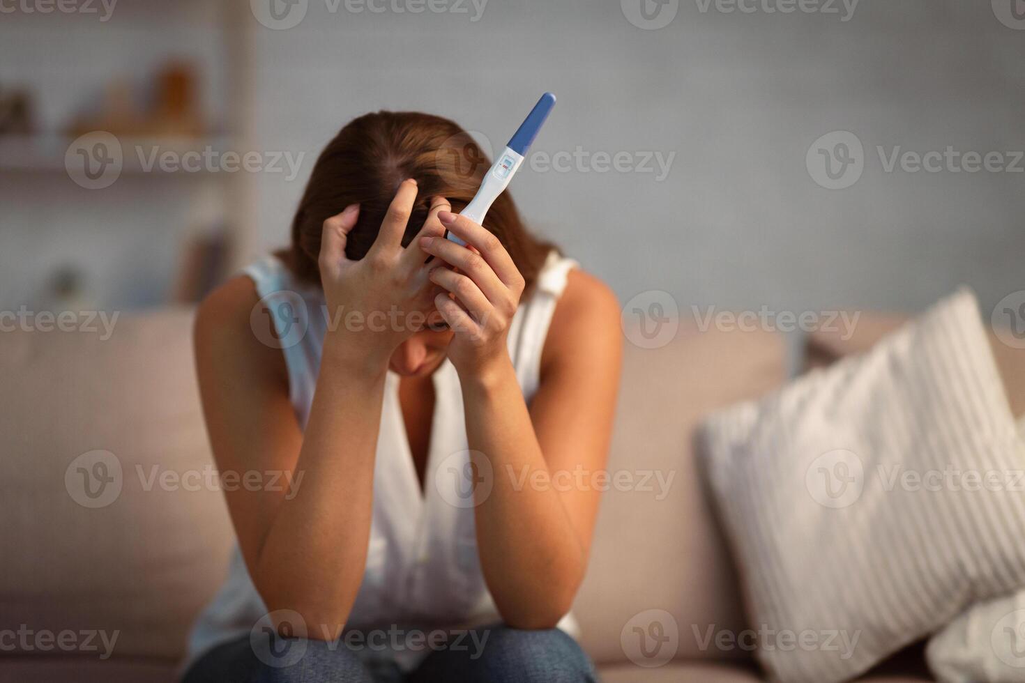 Unwanted pregnancy. Unrecognizable woman holding test covering face in despair sitting on sofa at home. Selective focus photo