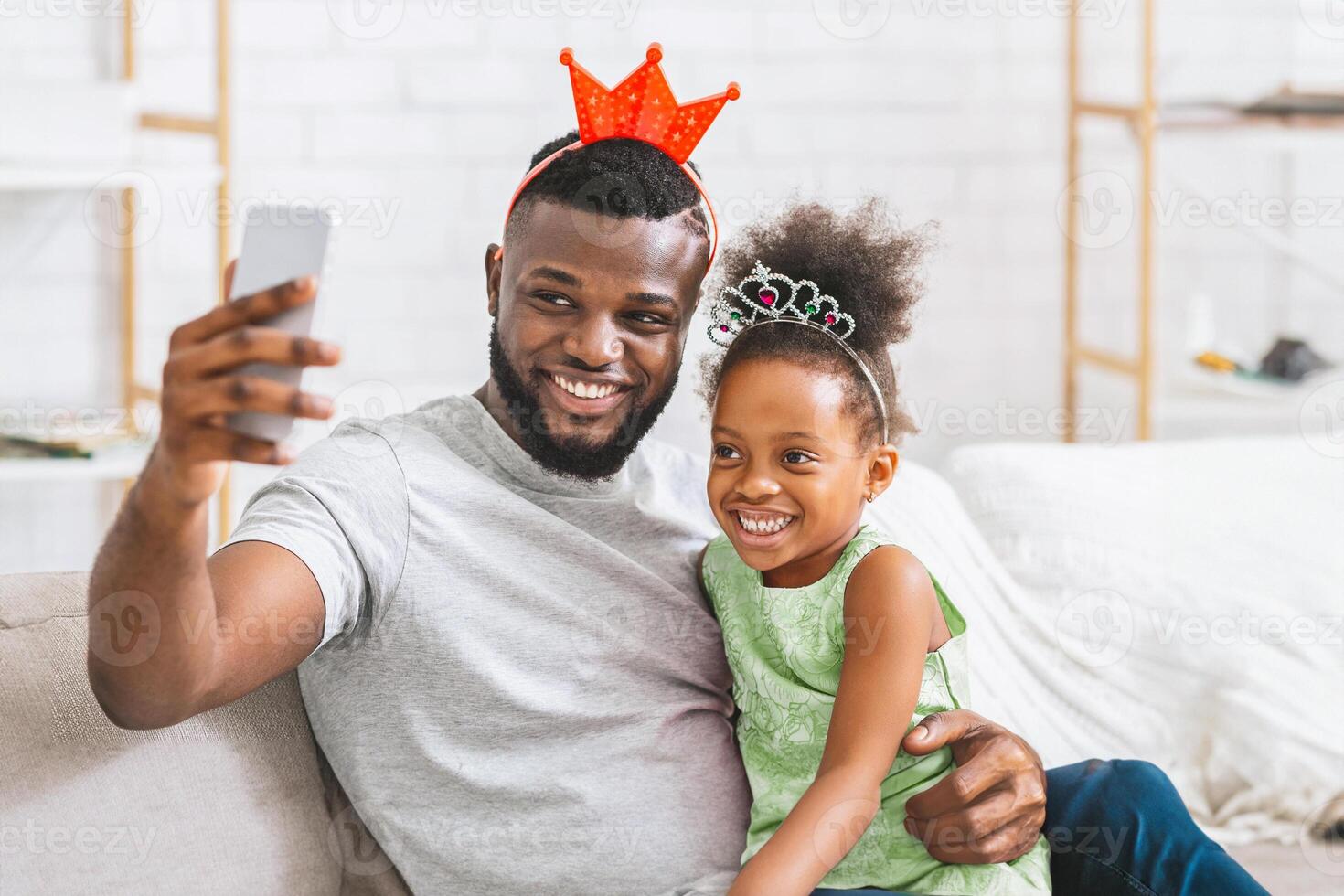 A happy black father and daughter, both wearing crowns, sit on the couch together and take a selfie with a smartphone. The father is smiling and holding the phone out in front of him. photo