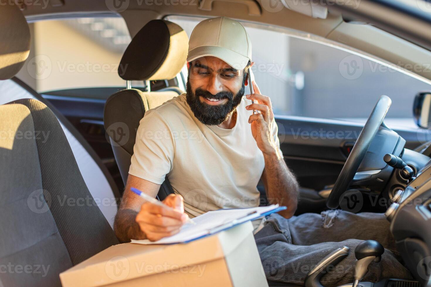 Indian delivery driver wearing a beige cap is seated inside his vehicle, talking on the phone and writing notes on a clipboard during the daytime. photo
