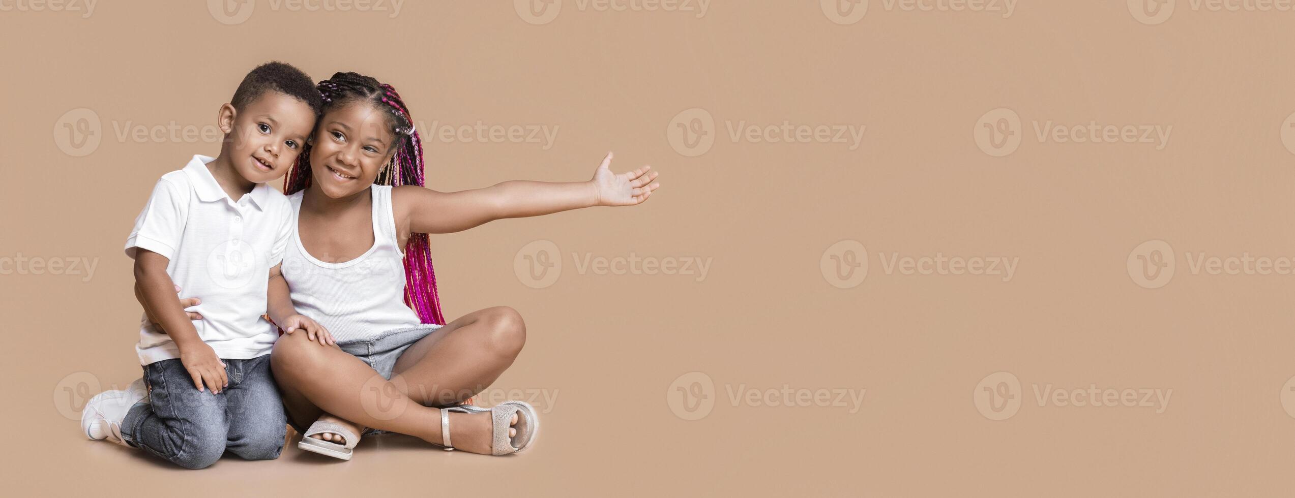 Adorable little african american siblings sitting on floor and cuddling, cute girl pointing aside with her open arm, yellow background with copy space photo