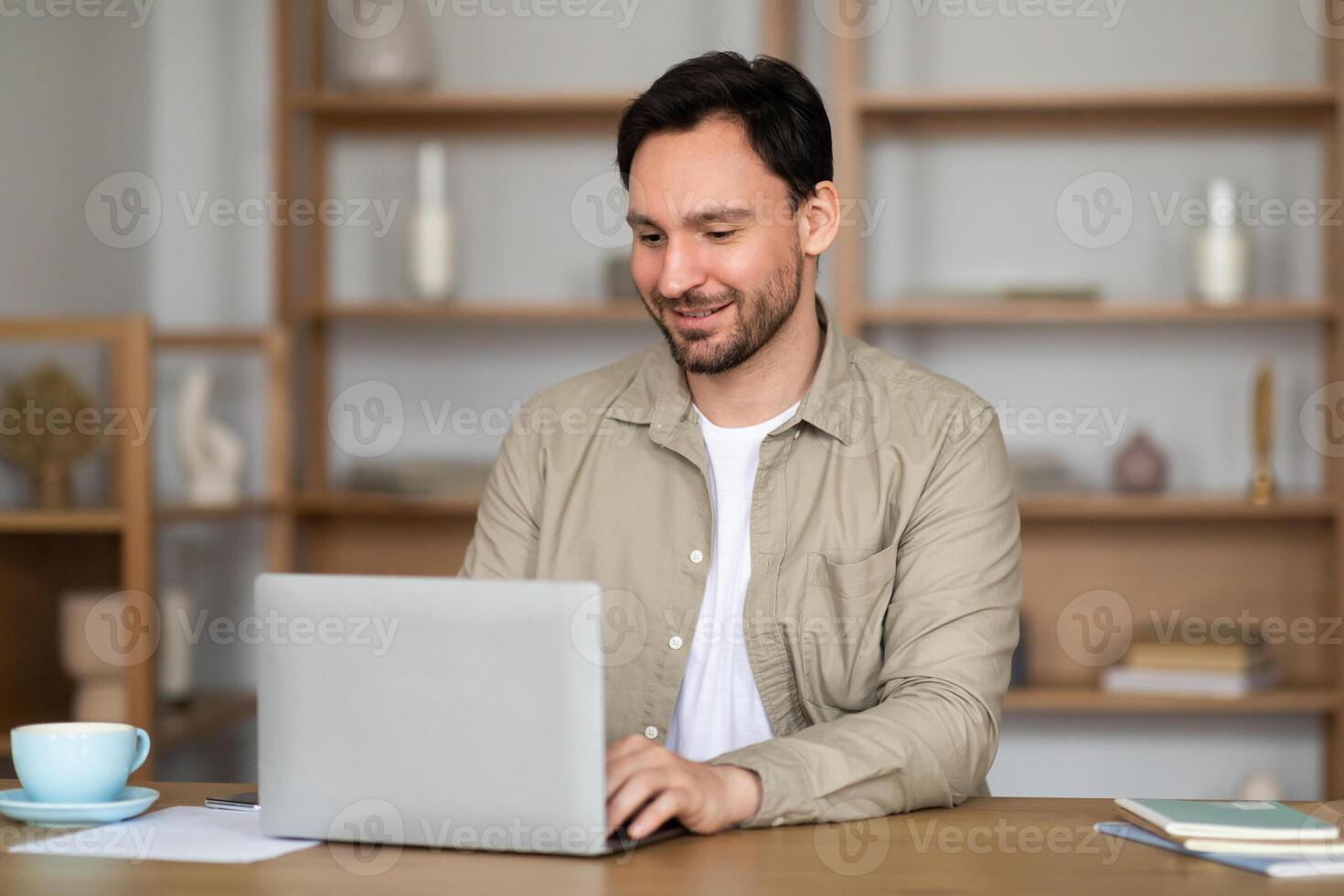 A man is seated at a table, focused on his laptop screen. Beside him, there is a steaming cup of coffee. The man appears engrossed in his work, typing on the laptop keyboard while sipping his coffee. photo