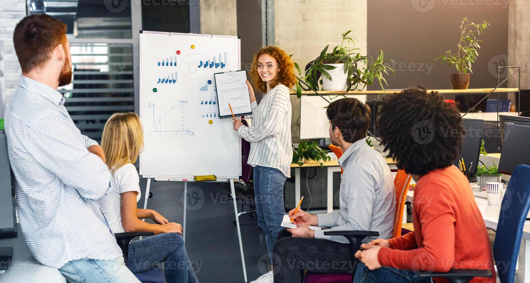 A confident young woman stands presenting a chart to a group of attentive colleagues during a collaborative meeting in a contemporary workspace photo
