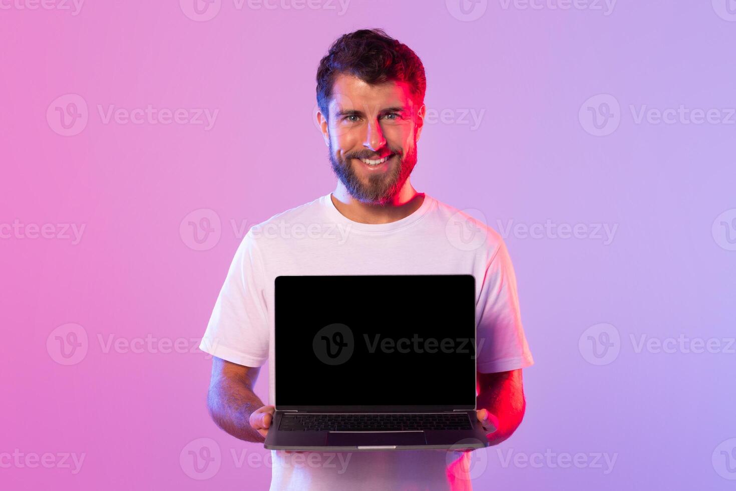 A young man in a white t-shirt smiles while holding an open laptop with black screen in a studio. The background features vibrant pink and purple lighting. photo