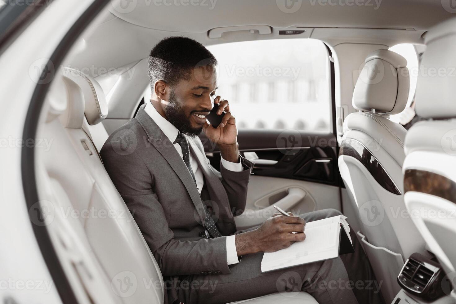 African American businessman in a suit sits in the backseat of a luxury car and makes notes on a notepad while speaking on his phone. He looks focused and professional. photo