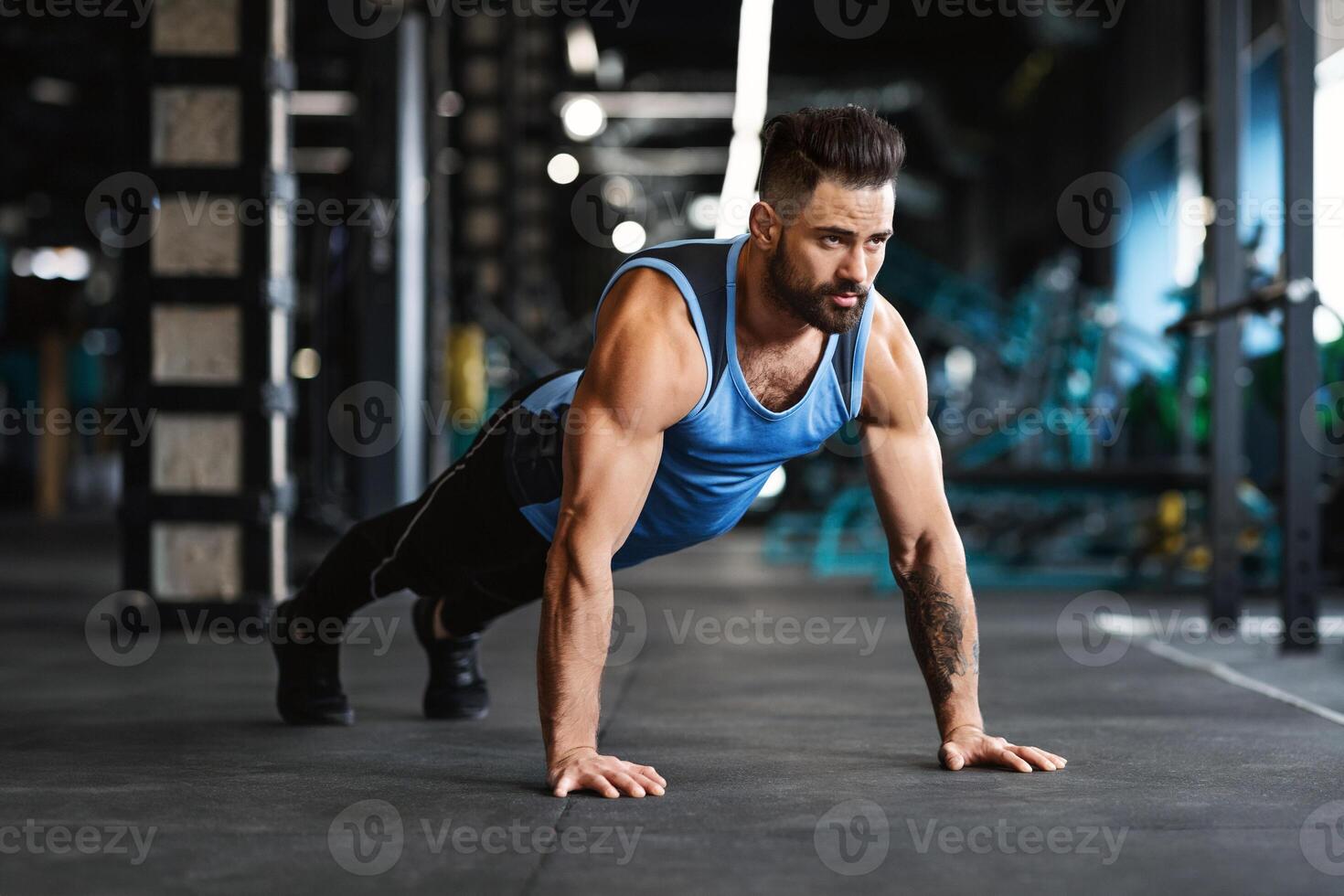 A man with a beard is doing push-ups in a gym. He is wearing a blue tank top and black pants. photo