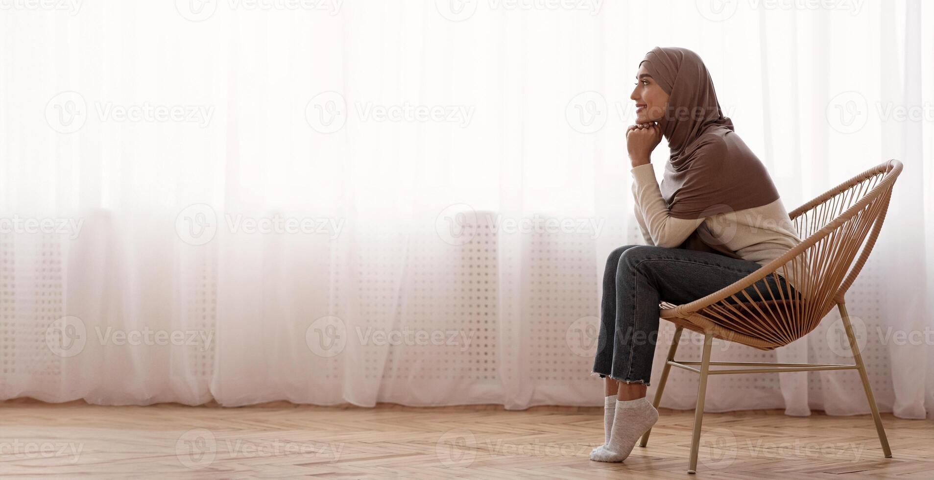 Morning Harmony. Muslim girl in hijab relaxing in wicker chair at home, resting head on hands and daydreaming against window, side view photo