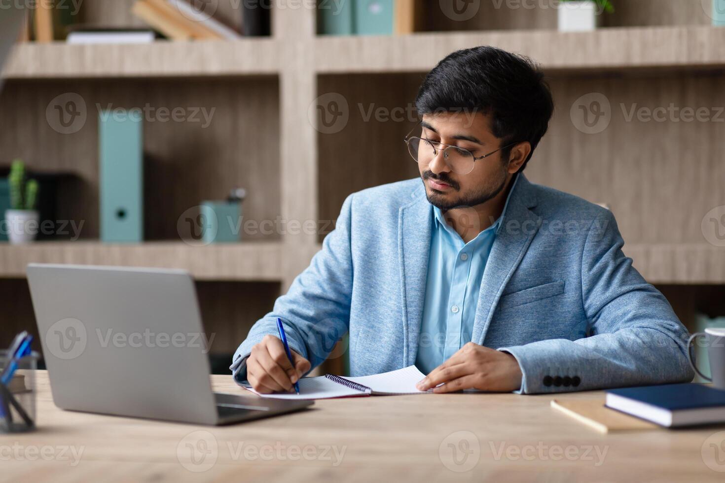 Indian man in a blue blazer and glasses sits at a wooden desk, writing in a notebook with a blue pen. His laptop is open in front of him, and there are a few other notebooks and a mug on the desk. photo