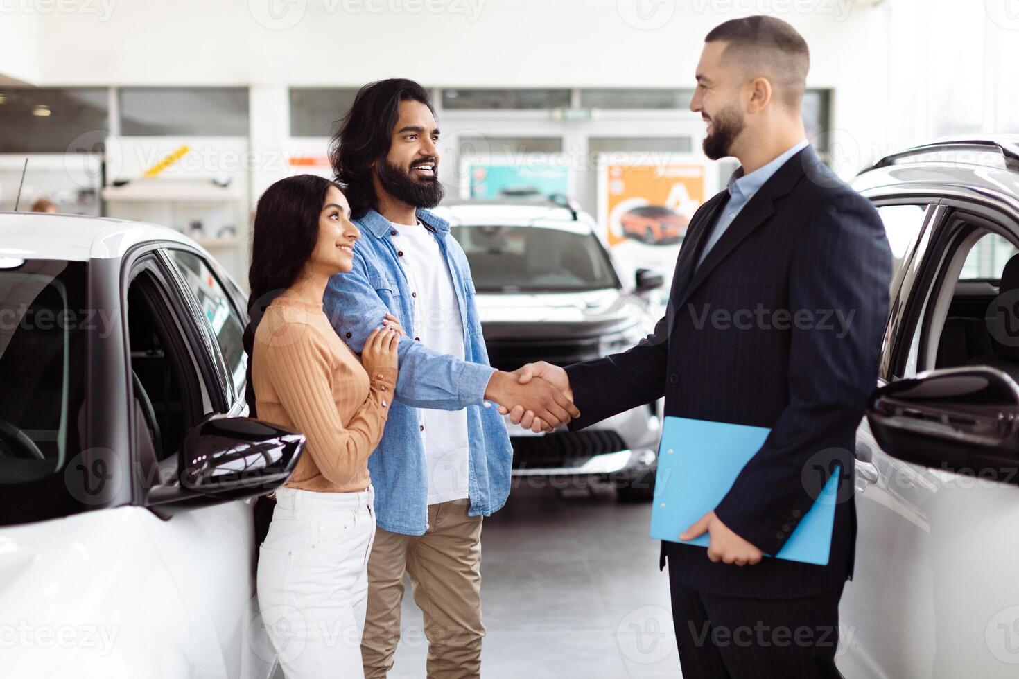 A young Indian couple is finalizing a deal with a car salesman inside a vehicle showroom. They seem pleased and are shaking hands with the agent, who is dressed in a business suit photo