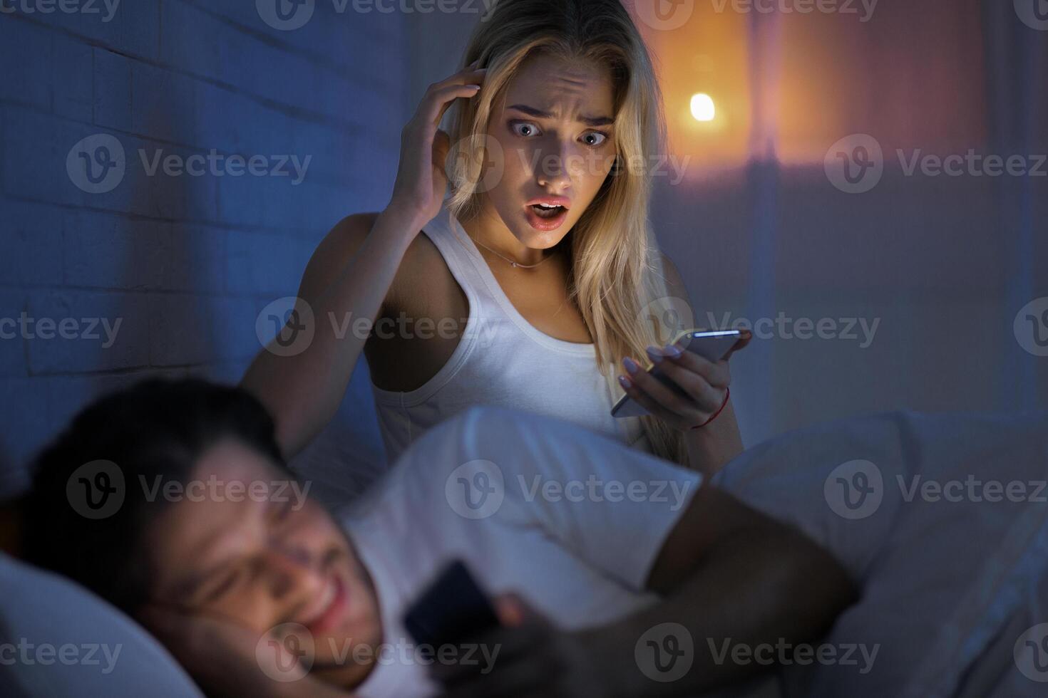 Online Affair. Shocked Woman Catching Boyfriend Cheater Chatting On Phone Lying In Bed Indoor. Low Light, Selective Focus photo