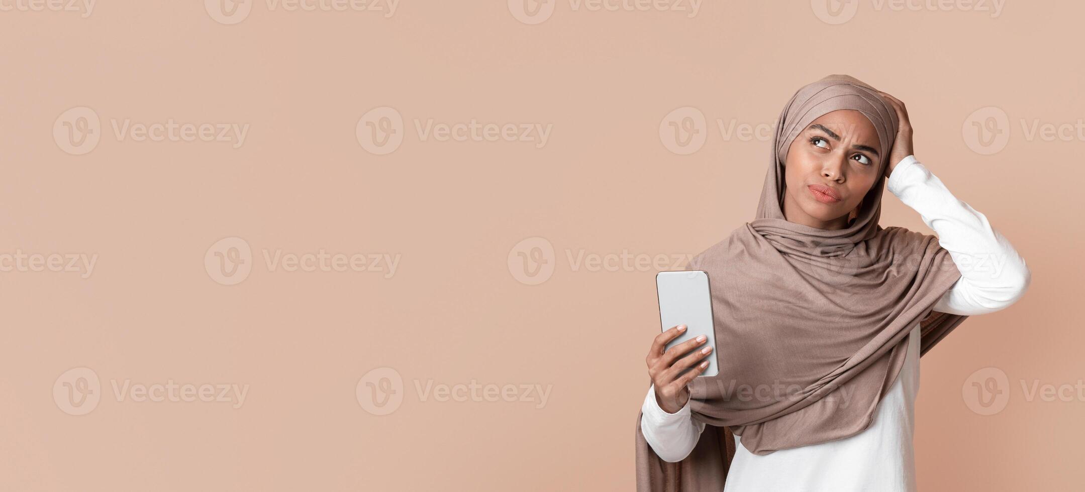 Forgot phone password. Thoughtful afro muslim girl holding smartphone and touching her head, not able to unblock her device, yellow blackground photo