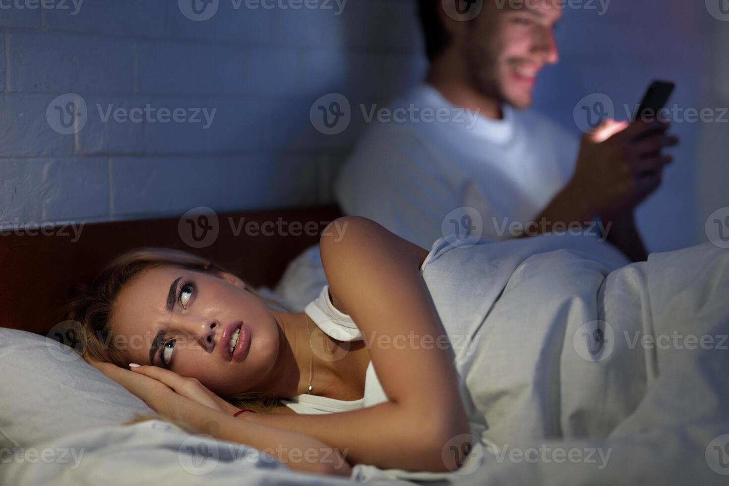Jealous Wife Suspecting Infidelity While Husband Texting On Mobile Phone Lying In Bed At Home At Night. Low Light photo