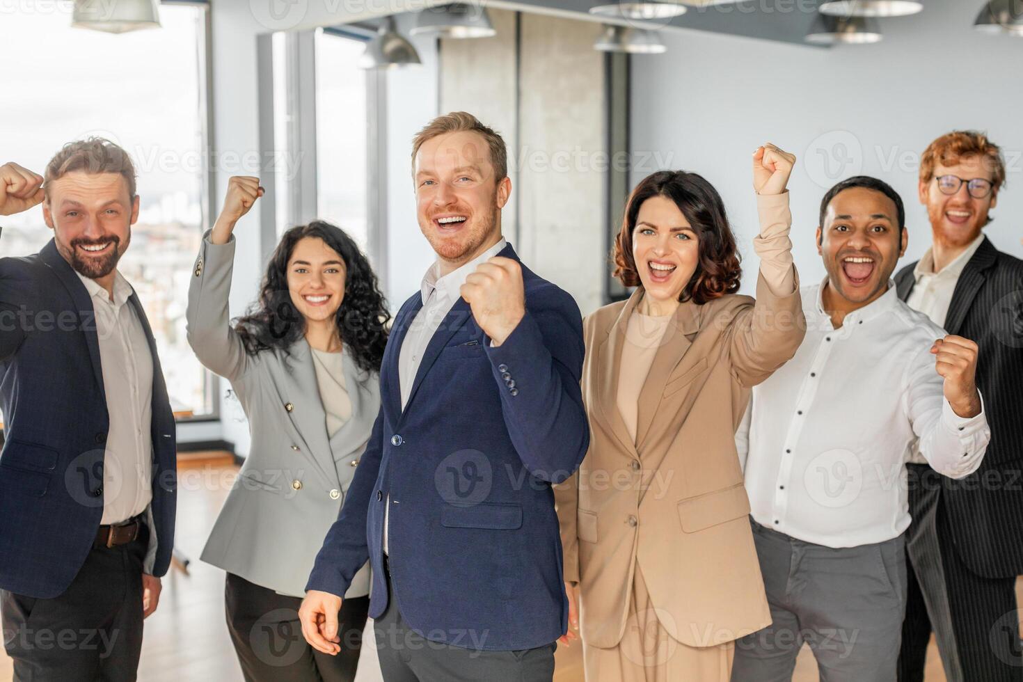 A diverse group of five business professionals stand in a modern office setting, with each person raising a fist in the air and smiling broadly, celebrating a successful achievement. photo