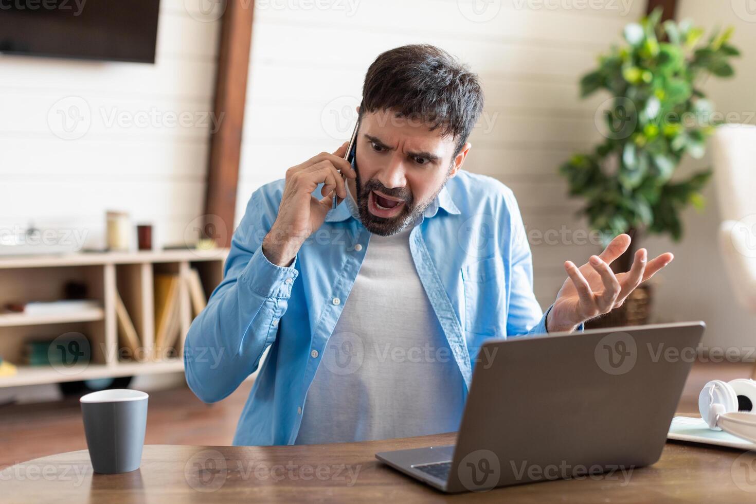 A man is seated at his work desk at home, expressing frustration while speaking on his mobile phone. He appears agitated, gesturing with his free hand photo