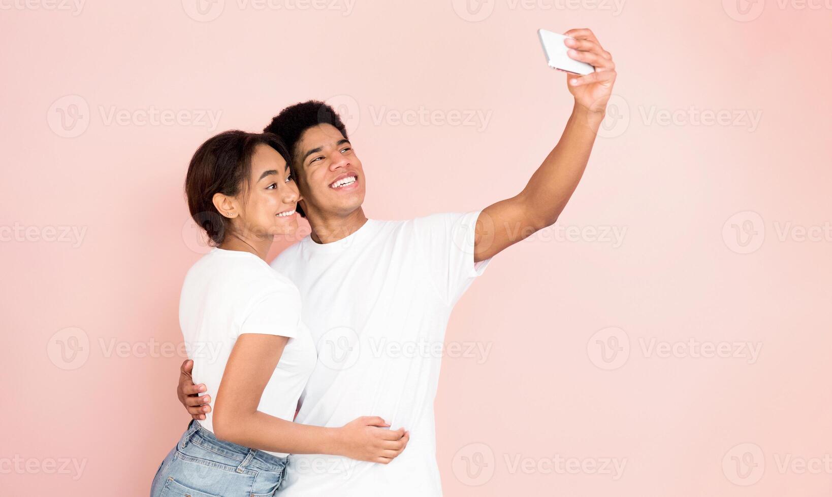 A young African American couple is seen smiling while taking a selfie against a vibrant pink background. The couple appears joyful and engaged in the act of capturing a moment together. photo
