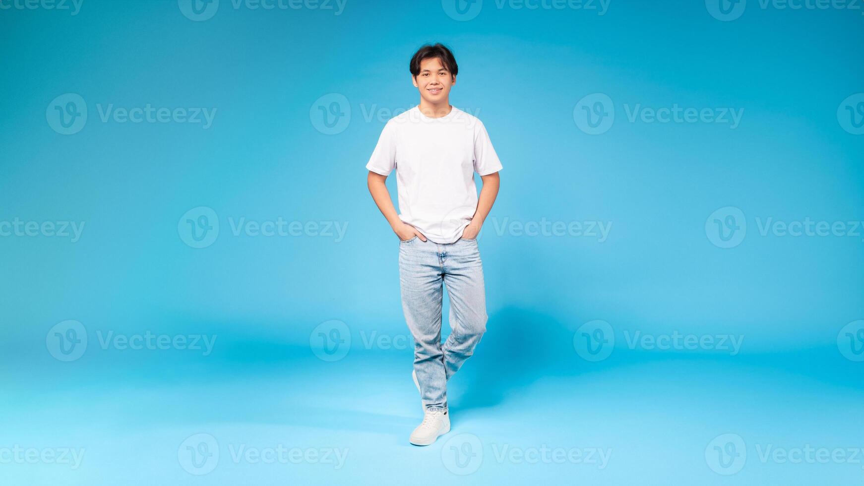 Asian guy in a white t-shirt is standing upright in front of a plain blue background. His posture is relaxed, with arms by his side and feet grounded on the floor. photo