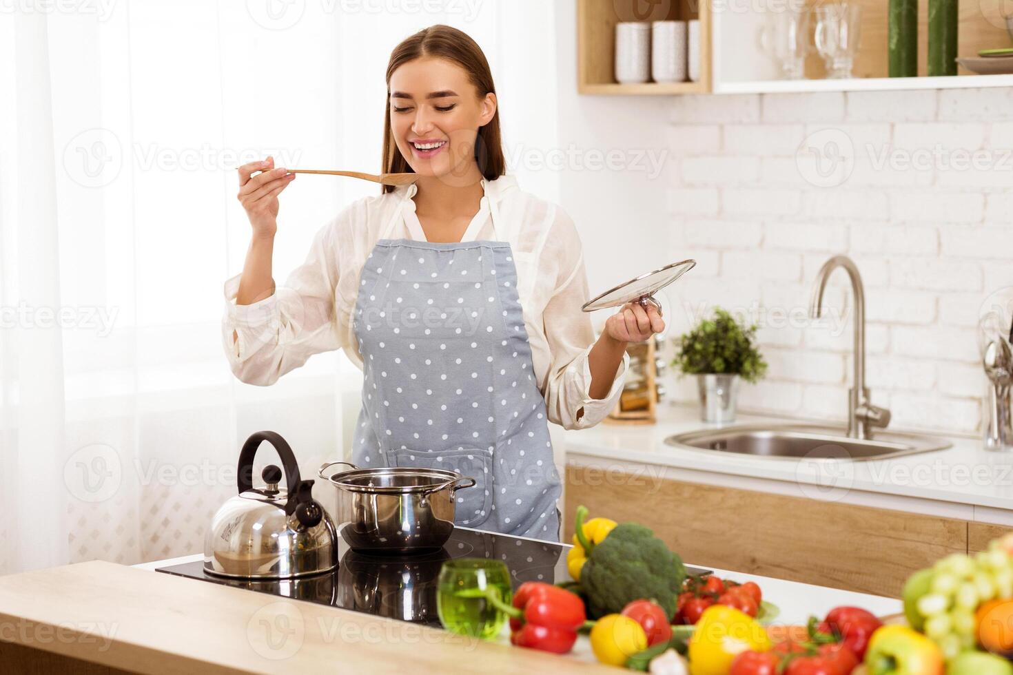 A happy woman in an apron tastes food while cooking in her kitchen. She is surrounded by fresh vegetables and cooking supplies. photo