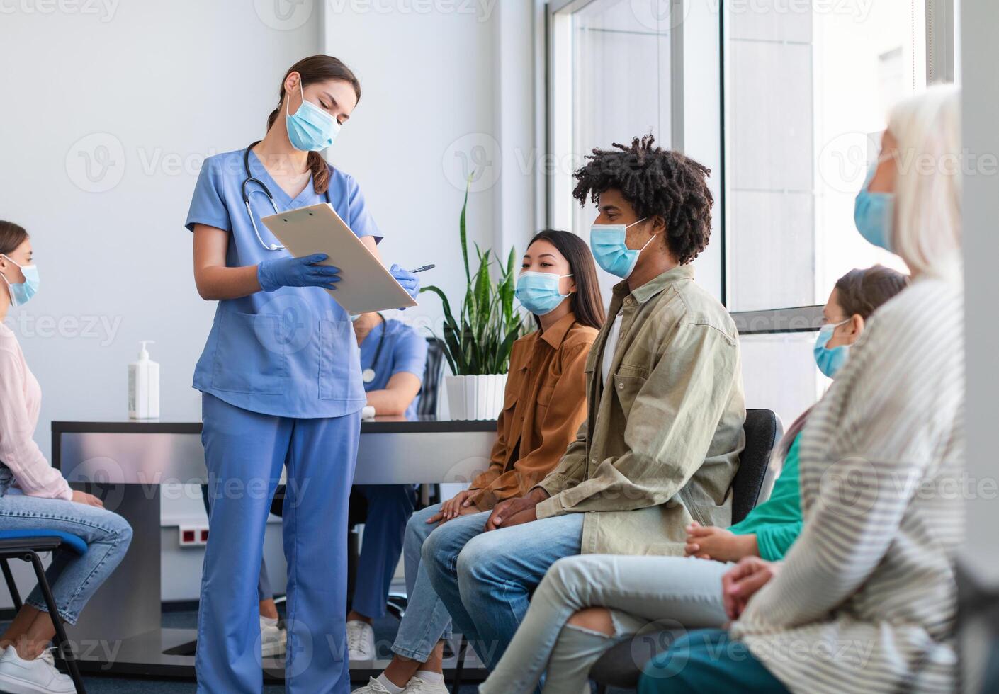 Coronavirus Vaccination Campaign Concept. Female doctor or nurse in uniform holding clipboard while talking to patients sitting and waiting in line at medical clinic, showing consent form to sign photo