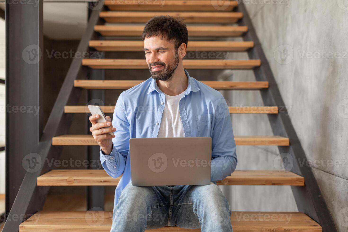 A man sits on wooden stairs in a modern interior. He is holding a smartphone in one hand and has a laptop on his lap. The man is smiling as he looks down at his phone. photo