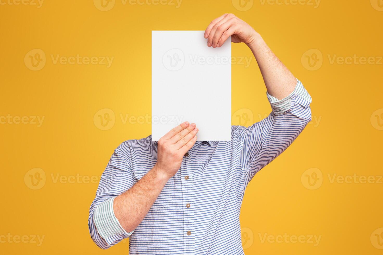 A man holding a blank white piece of paper over his face in the air against a plain yellow studio background, mockup, copy space photo