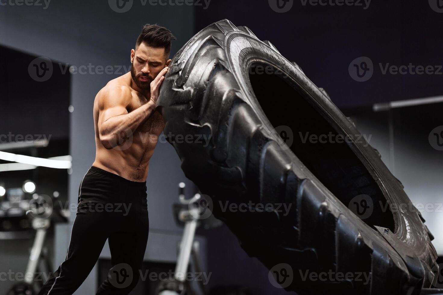A shirtless man with a muscular build lifts a heavy tire during a workout session at the gym. photo