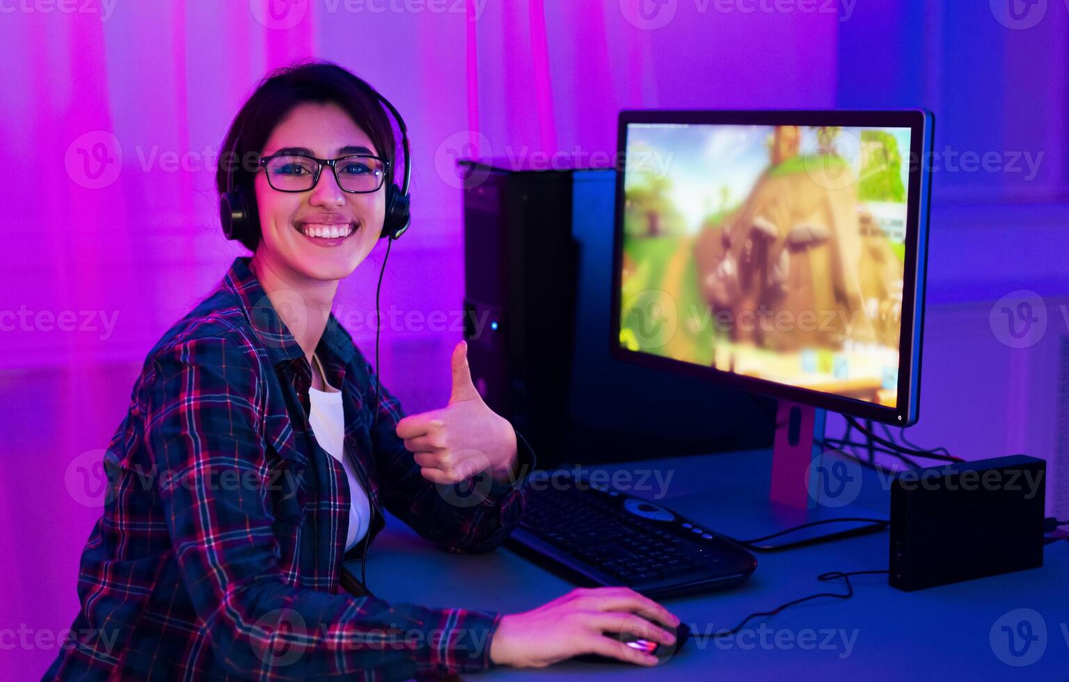 A woman in a plaid shirt sits at a computer desk with a gaming monitor displaying an animated scene. She wears headphones and smiles at the camera, giving a thumbs up photo