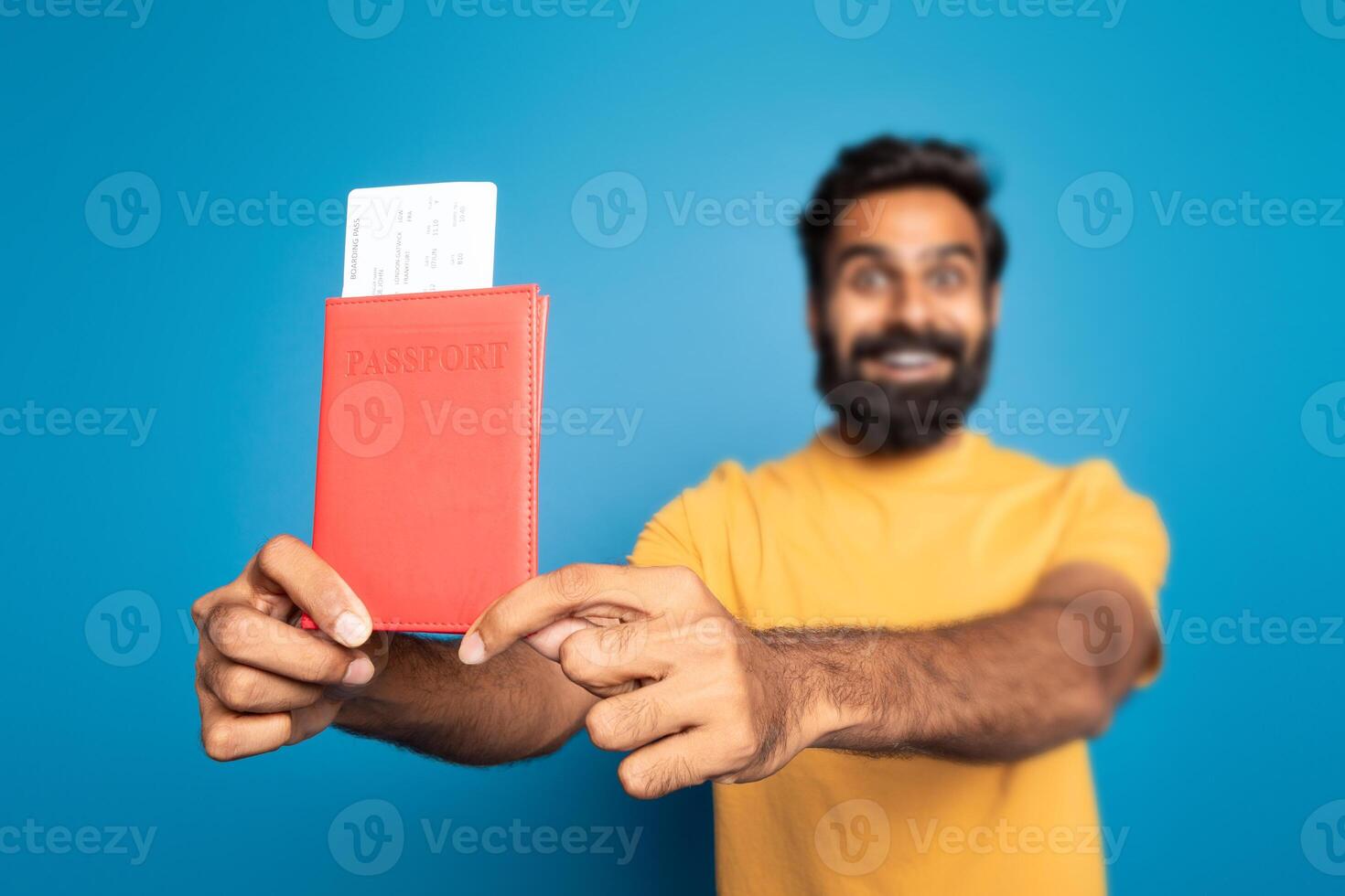 A bearded hindu man in a yellow shirt enthusiastically holds up a red passport with a boarding pass sticking out, blue background emphasizes the subject, likely indicating travel or vacation plans. photo