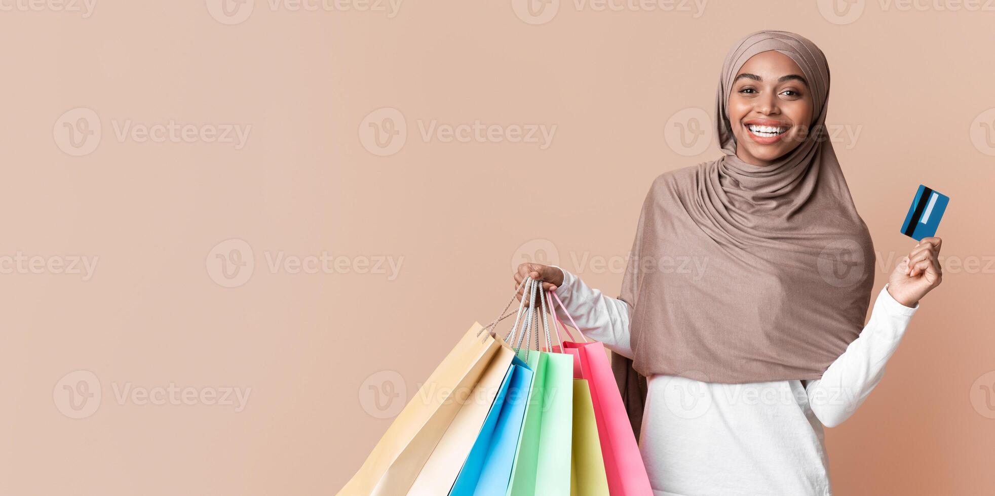 Cashback bonus concept. Happy black muslim girl in hijab holding credit card and colourful shopping bags over yellow background, free space photo