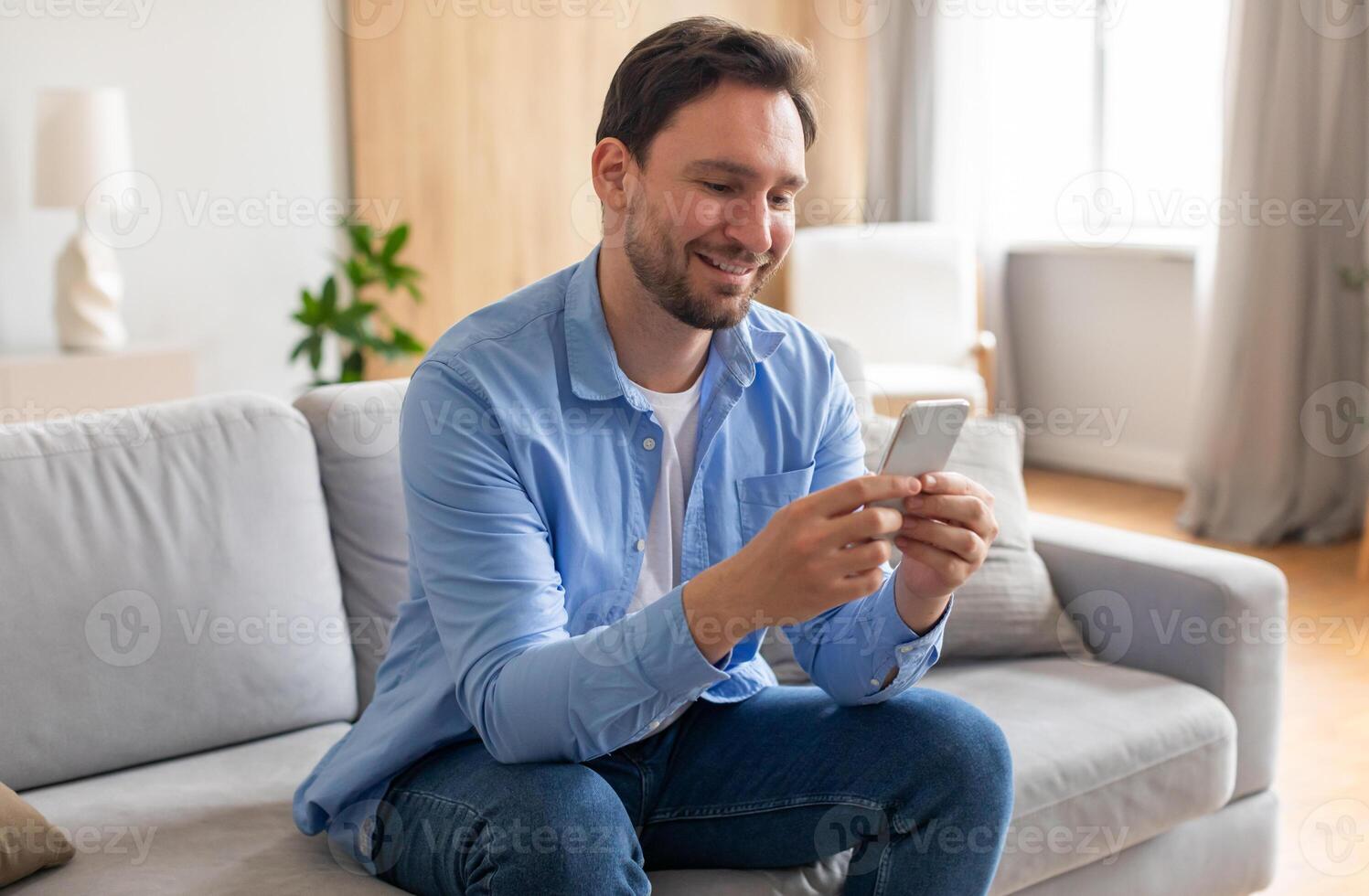 A bearded man is seated on a couch, engrossed in his smartphone screen. He appears focused as he scrolls and interacts with the device, surrounded by a living room setting. photo