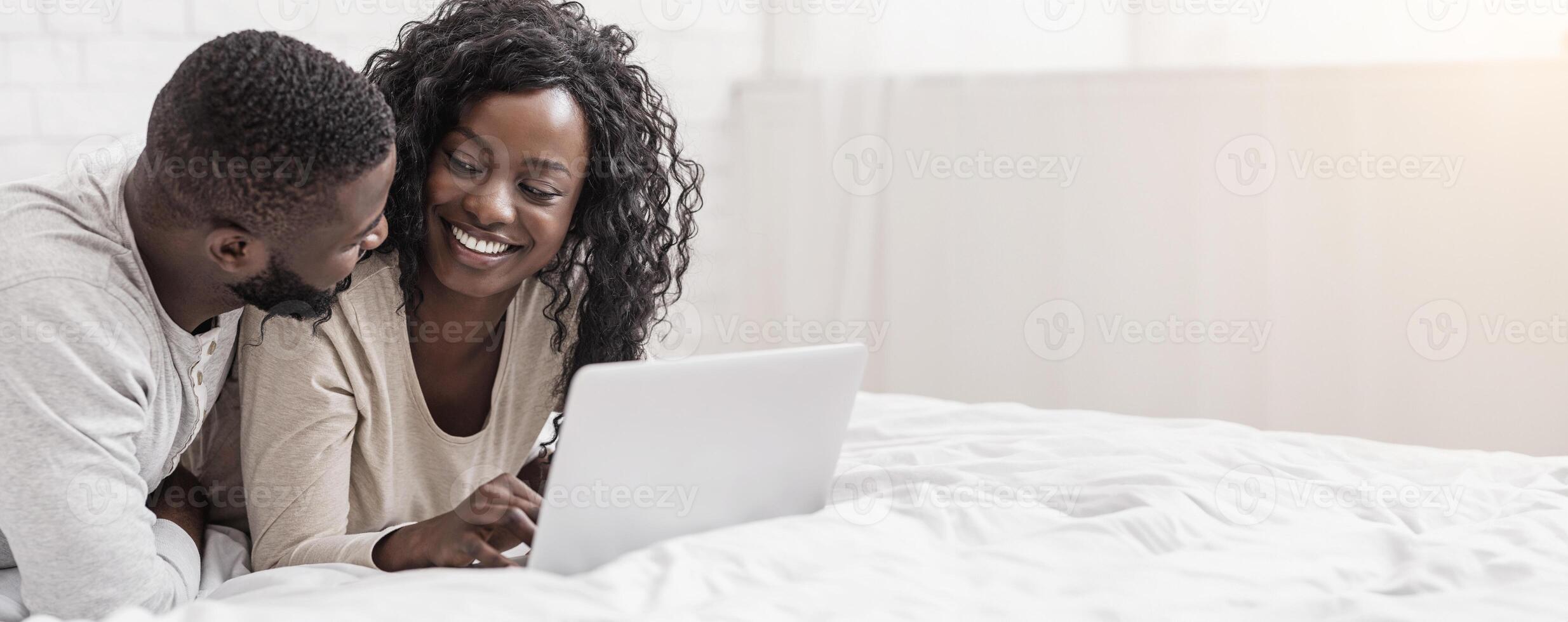 Smiling young afro couple using laptop in bed at home, browsing social media or surfing internet, free space photo