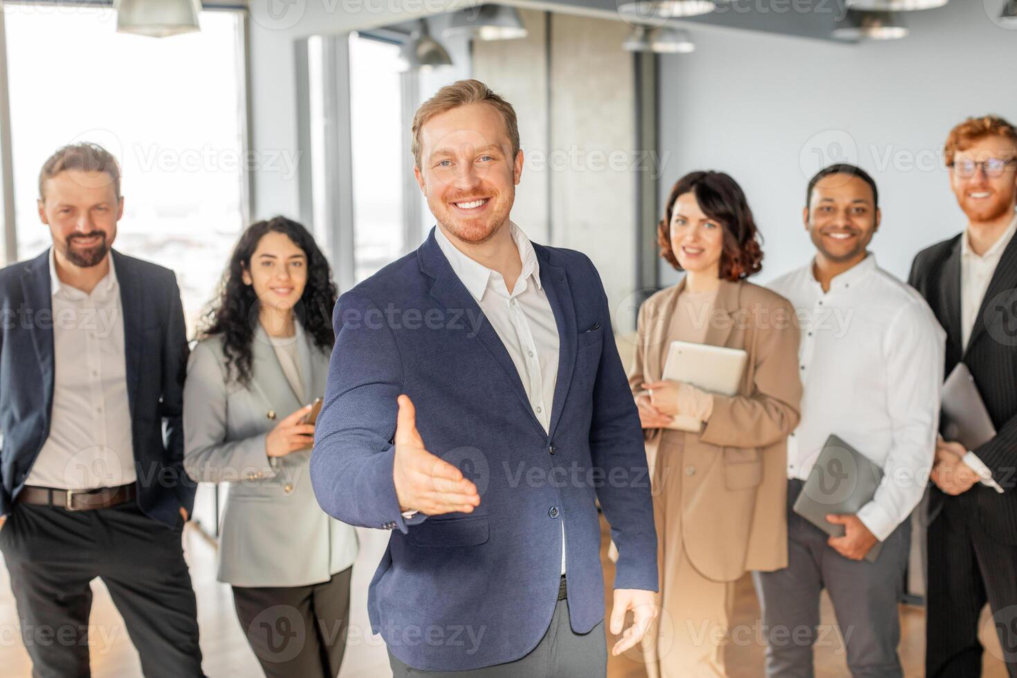 A group of diverse business professionals are standing in a modern office, with one man extending his hand for a handshake. The others are smiling and looking at the camera. photo