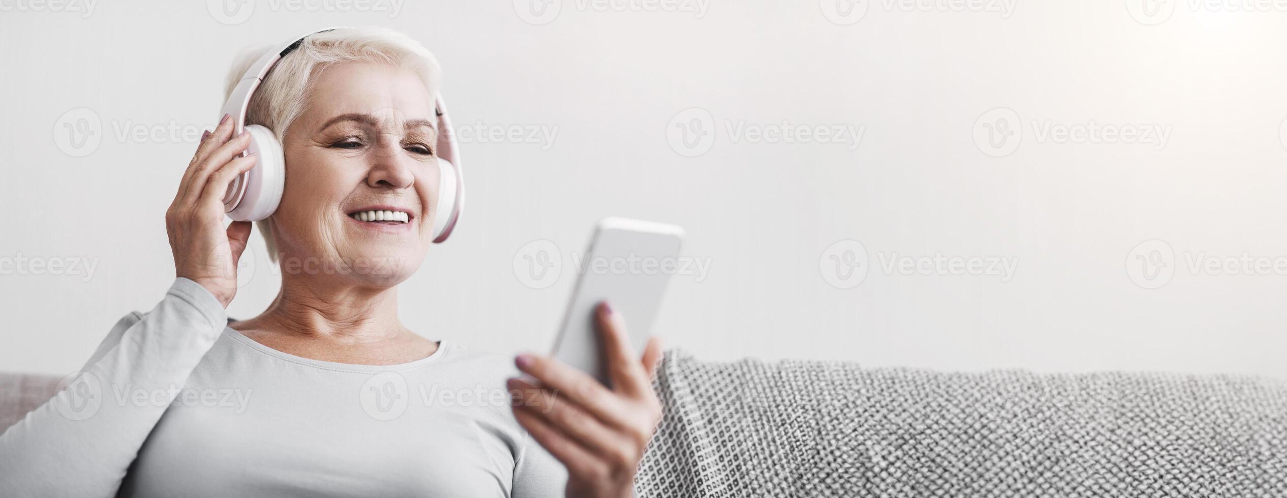 Senior woman with wireless headset enjoying music and watching videos on smartphone, home interior photo