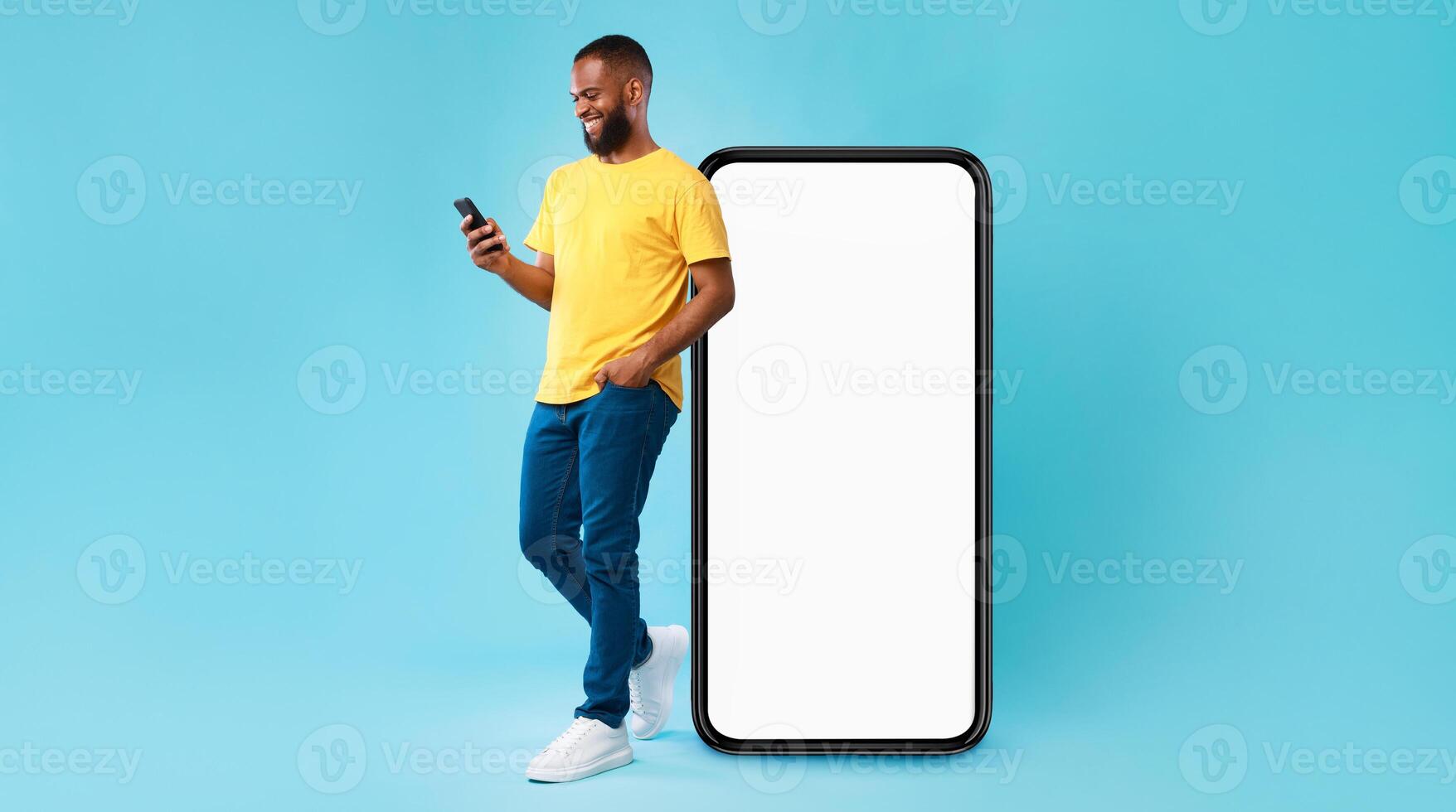 Handsome young Afro man using mobile phone while standing near huge smartphone with empty screen over blue studio background. Mockup for website or application, space for advertisement photo