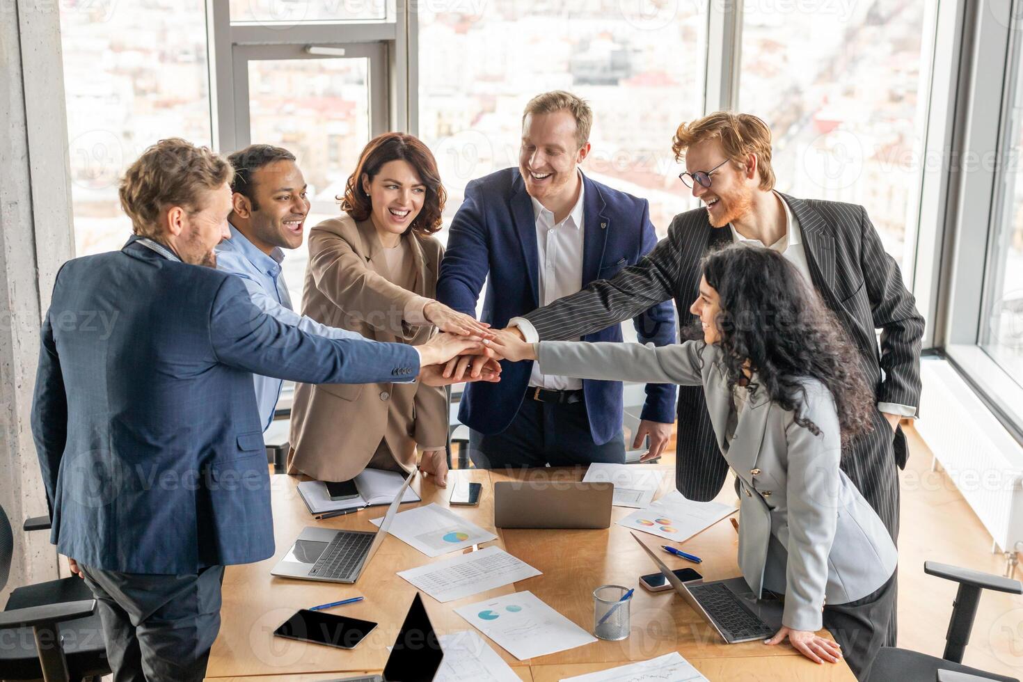 A diverse group of six business professionals stand around a conference table, their hands stacked high in the air as they celebrate a successful project. photo