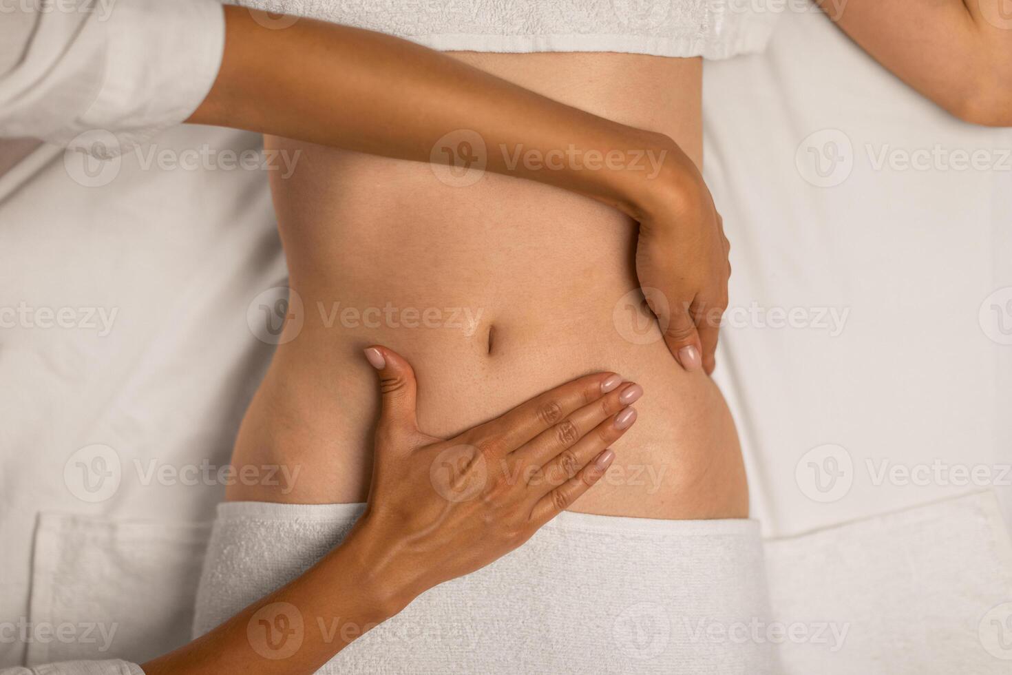 A physical therapists hands are assisting a patient by guiding and supporting their abdomen during rehabilitation exercises. The patient is lying down, wearing a white top and partially visible pants photo