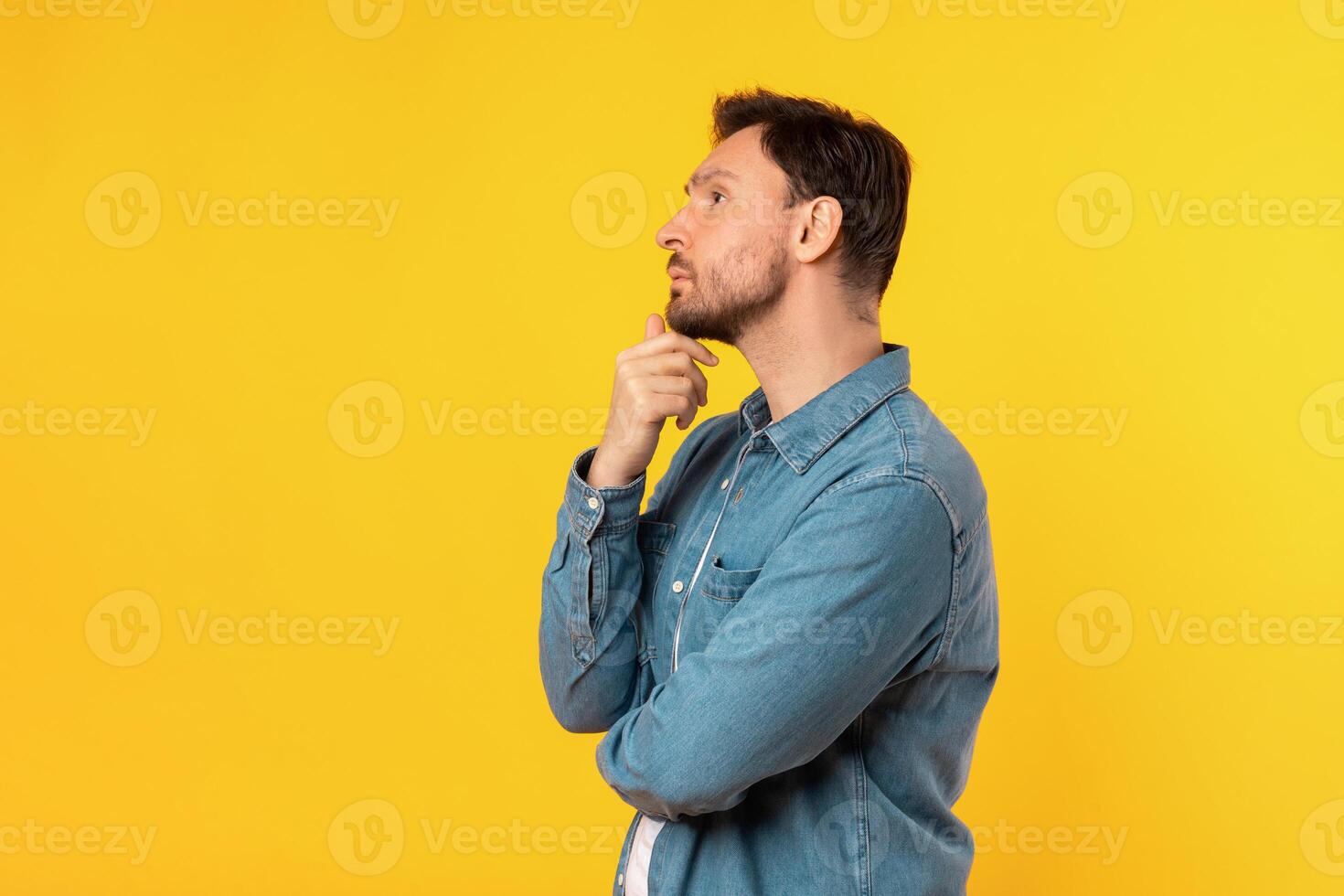 A man standing upright in front of a solid yellow background. The man is facing forward with a neutral expression, touching his chin and looking at copy space photo