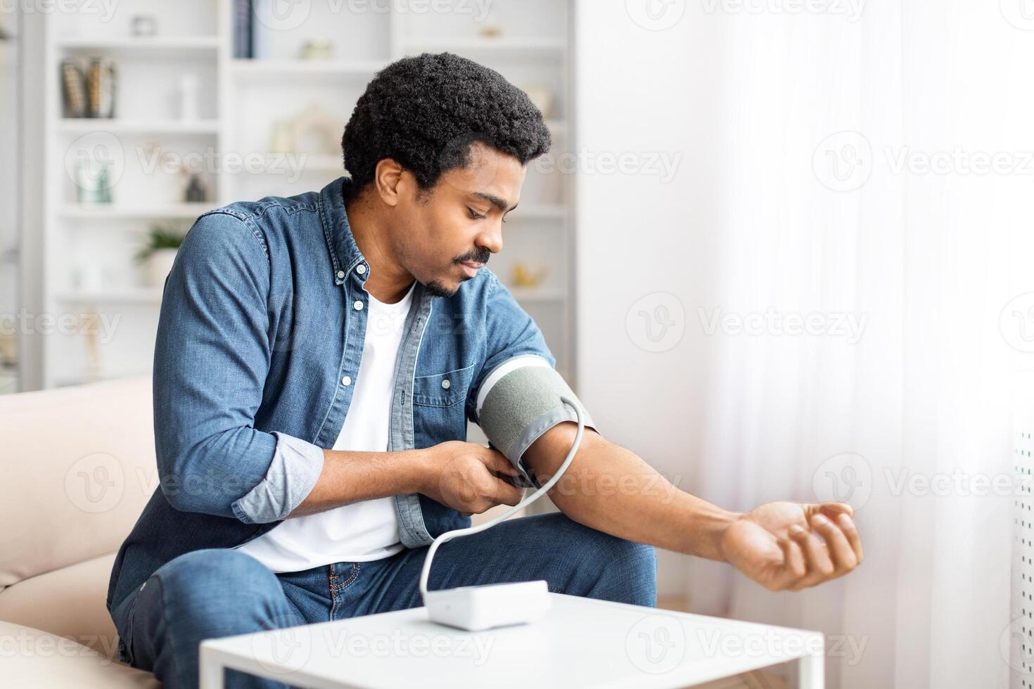 A focused young African American man sits comfortably on a couch while he wraps a blood pressure cuff around his upper arm, monitoring his blood pressure in the relaxed atmosphere of his living room. photo