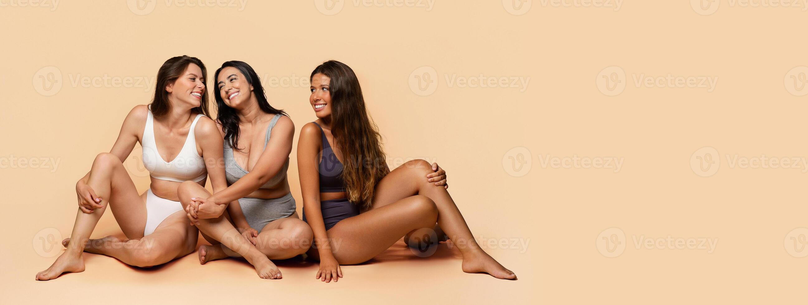 Three glad diverse millennial women in undergarments sit on floor, enjoy body positivity, beauty care, have fun, isolated on light studio background, full length. Friendship, confidence, copy space photo