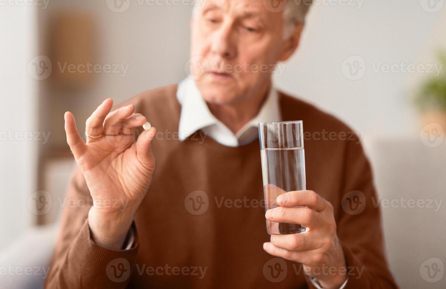 Medication Therapy. Senior Man Holding A Pill And Glass Of Water Sitting On Sofa At Home, Selective Focus photo