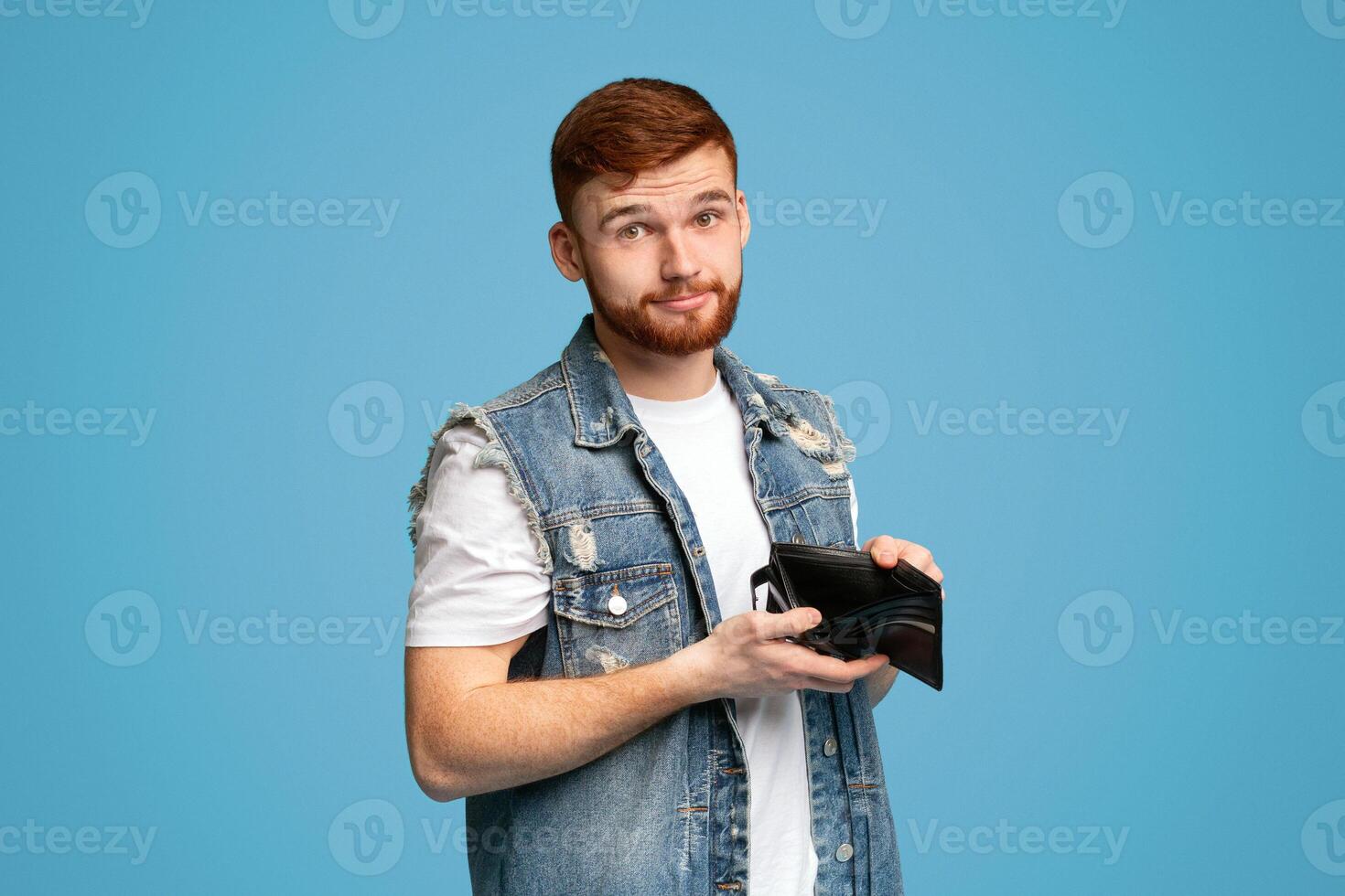 No money for living. Sad millennial man showing his empty wallet to camera on blue background, copy space photo