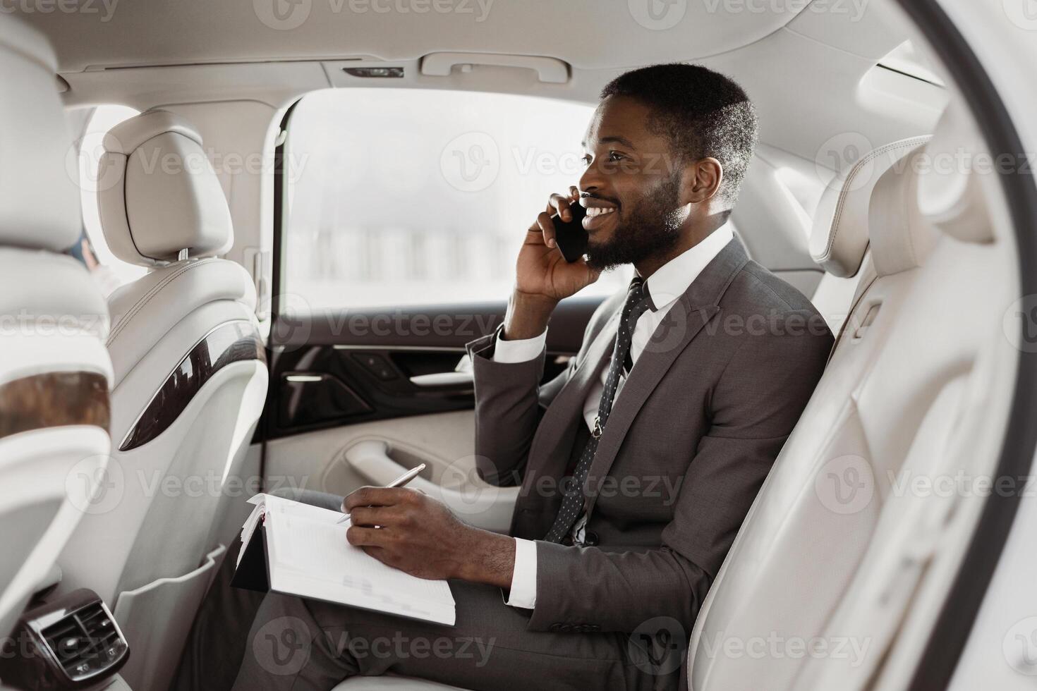 Black businessman is sitting in the backseat of a car, taking notes on a notepad while talking on a phone. He is wearing a suit and tie, and he has a serious expression on his face. photo