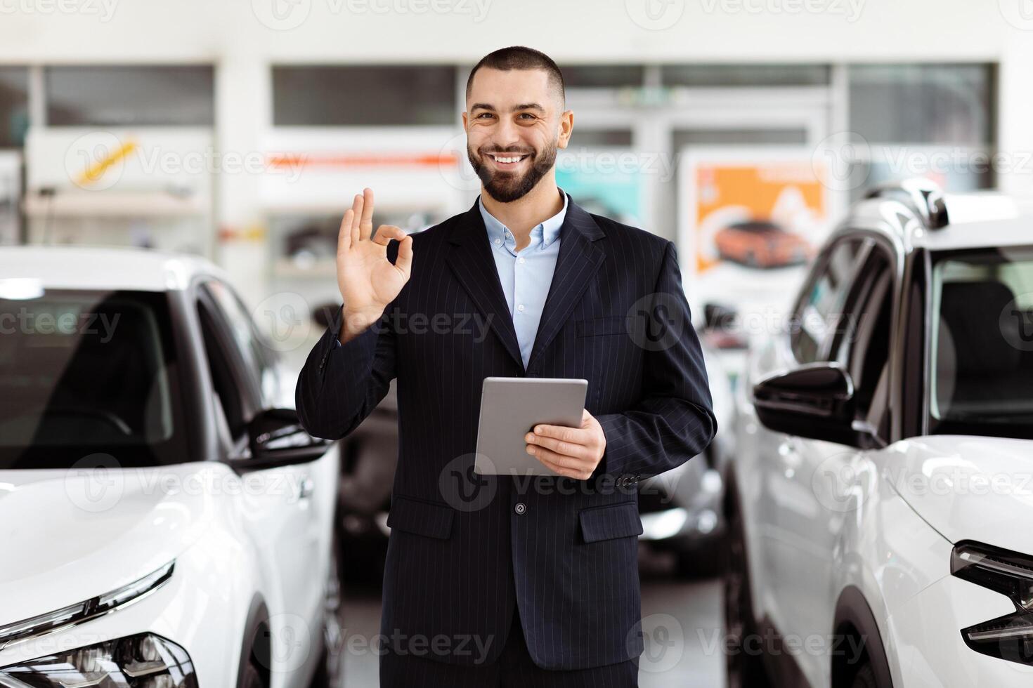 A man standing in front of a car, holding a tablet in his hand. He appears to be inspecting something on the device, with the car parked behind him. photo