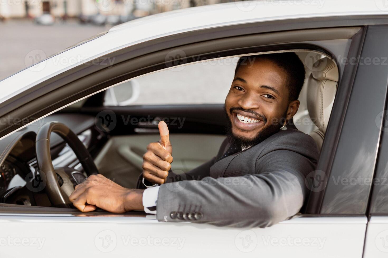 African American man in a suit smiles and gives a thumbs up as he sits in the drivers seat of a white car. photo